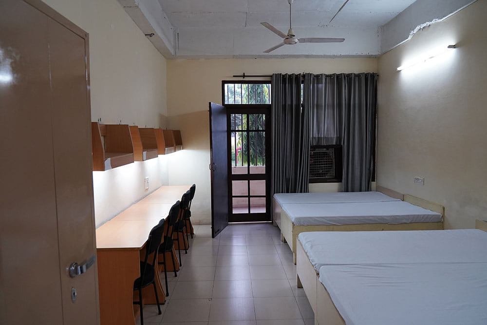 hostel room with two single beds, study desks and chairs, wall shelves, a ceiling fan, and a door opening to a small outdoor area